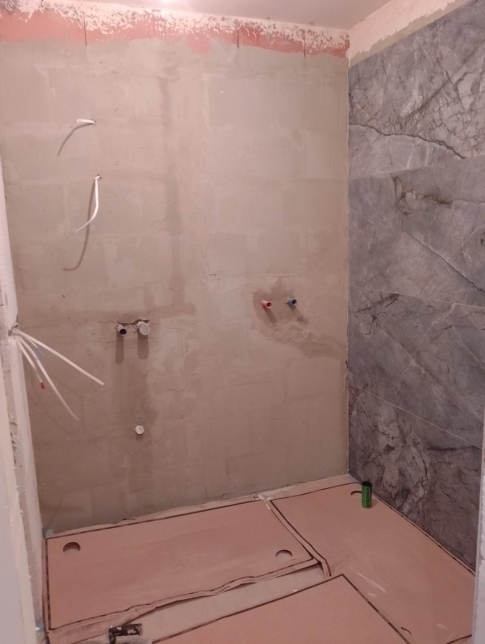 Łazienka w trakcie remontu: przygotowana ściana pod glazurę, częściowo ułożone płyty grzewcze podłogowe, widoczne rury instalacji wodnej.