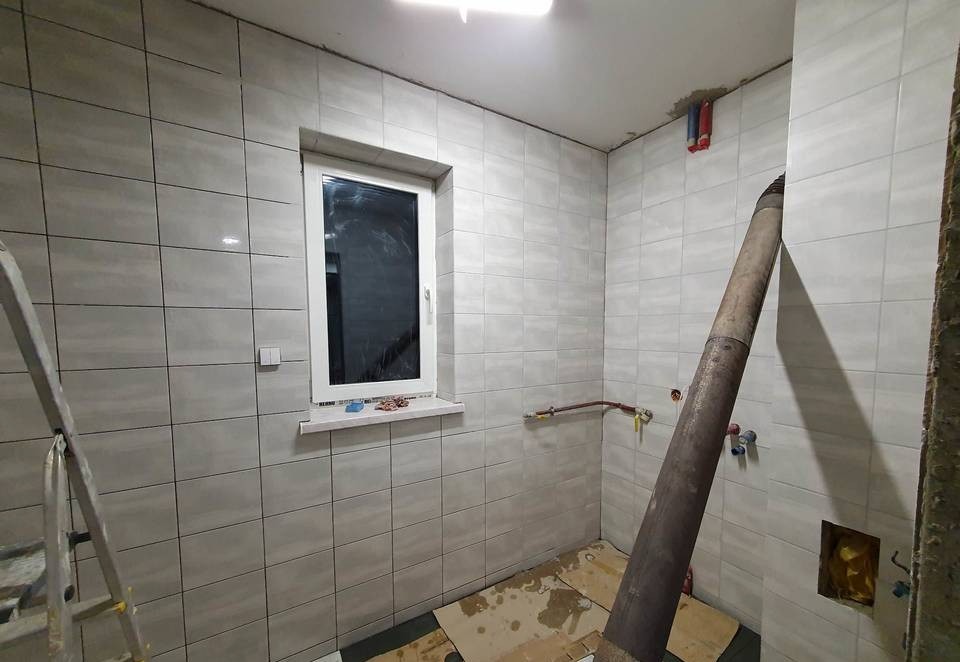 Łazienka w trakcie remontu: szare płytki ceramiczne na ścianach, okno z białymi ramami, widoczne instalacje hydrauliczne i rura wentylacyjna, podłoga zabezpieczona kartonem, drabina aluminiowa...