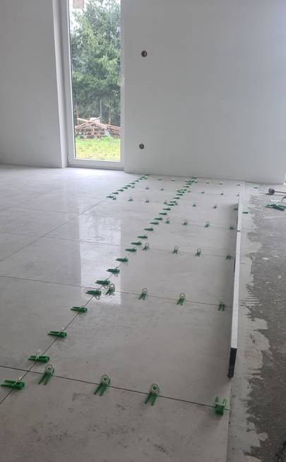 Układanie jasnych płytek podłogowych z użyciem zielonych klipsów poziomujących i sznurka w nowym, jasnym pomieszczeniu z dużym oknem.