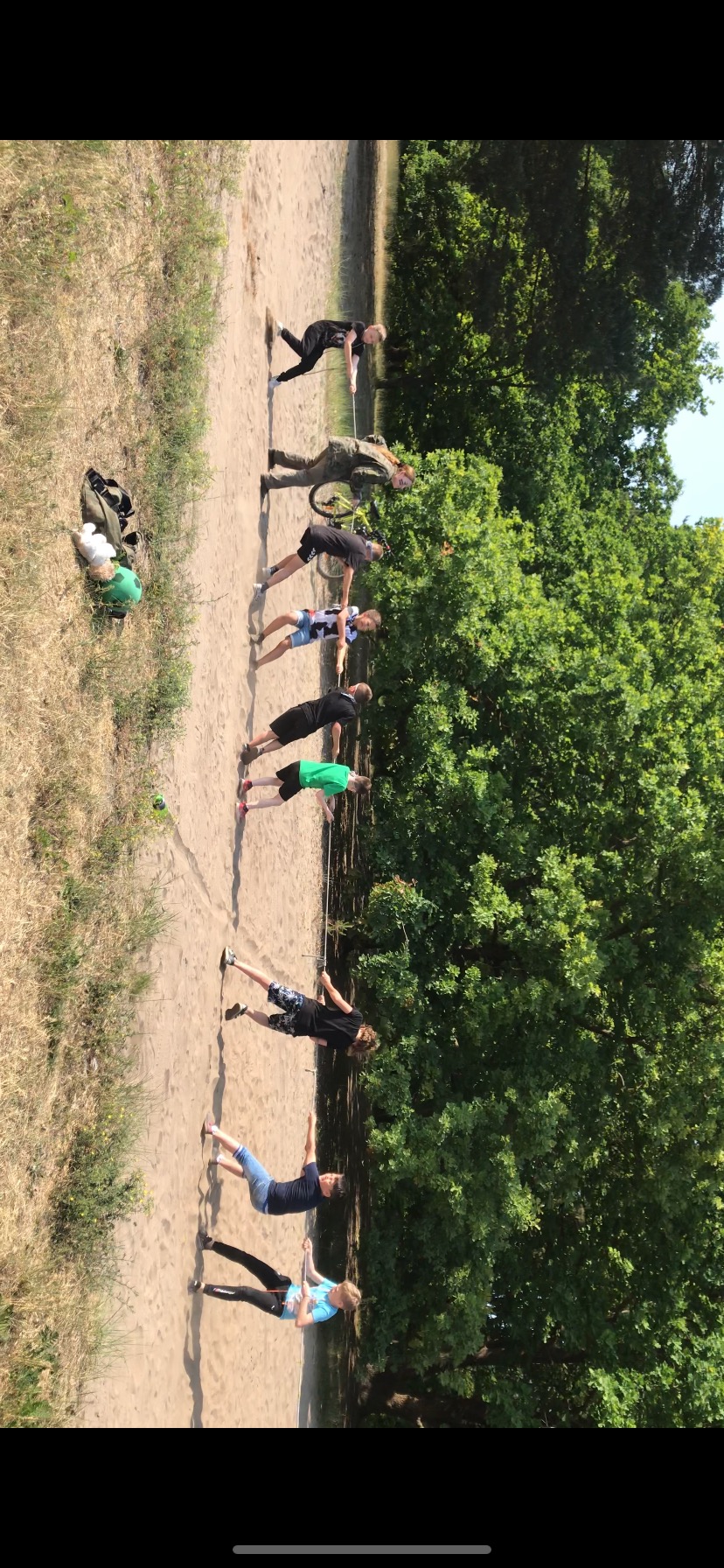 Grupa dzieci na piasku, trzymających się liny podczas zabawy na świeżym powietrzu, obok leżą plecak, piłka i pluszaki.