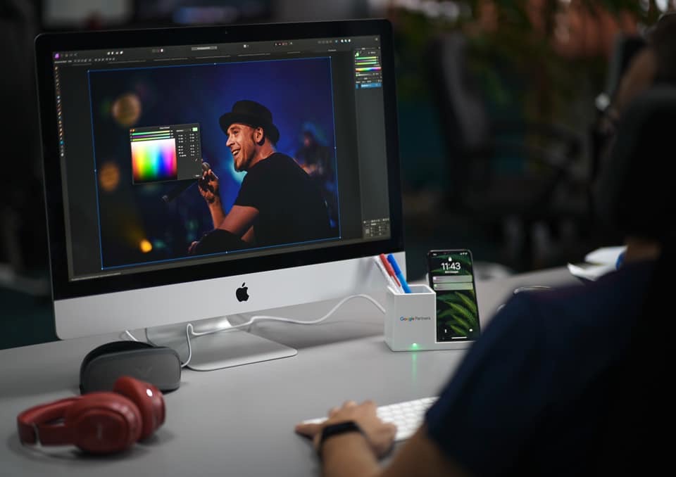 Grafik komputerowy edytuje zdjęcie piosenkarza w kapeluszu, używając palety kolorów na ekranie iMac. Na biurku słuchawki, telefon i organizer z długopisami.