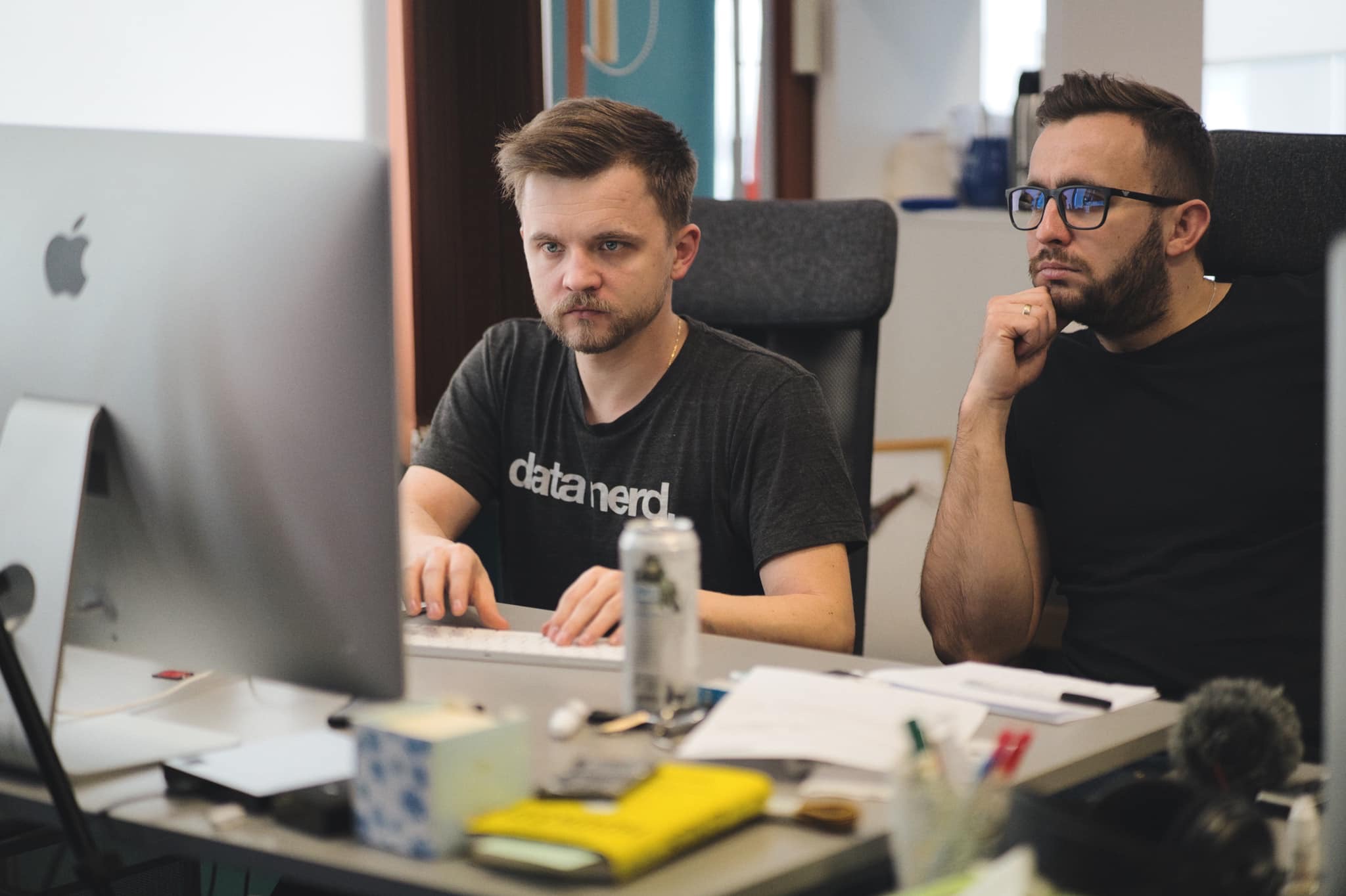 Dwóch mężczyzn w biurze skupionych na pracy przed dużym monitorem Apple, jeden pisze na klawiaturze, drugi z brodą i okularami zamyślony, oparty podbródek na dłoni.