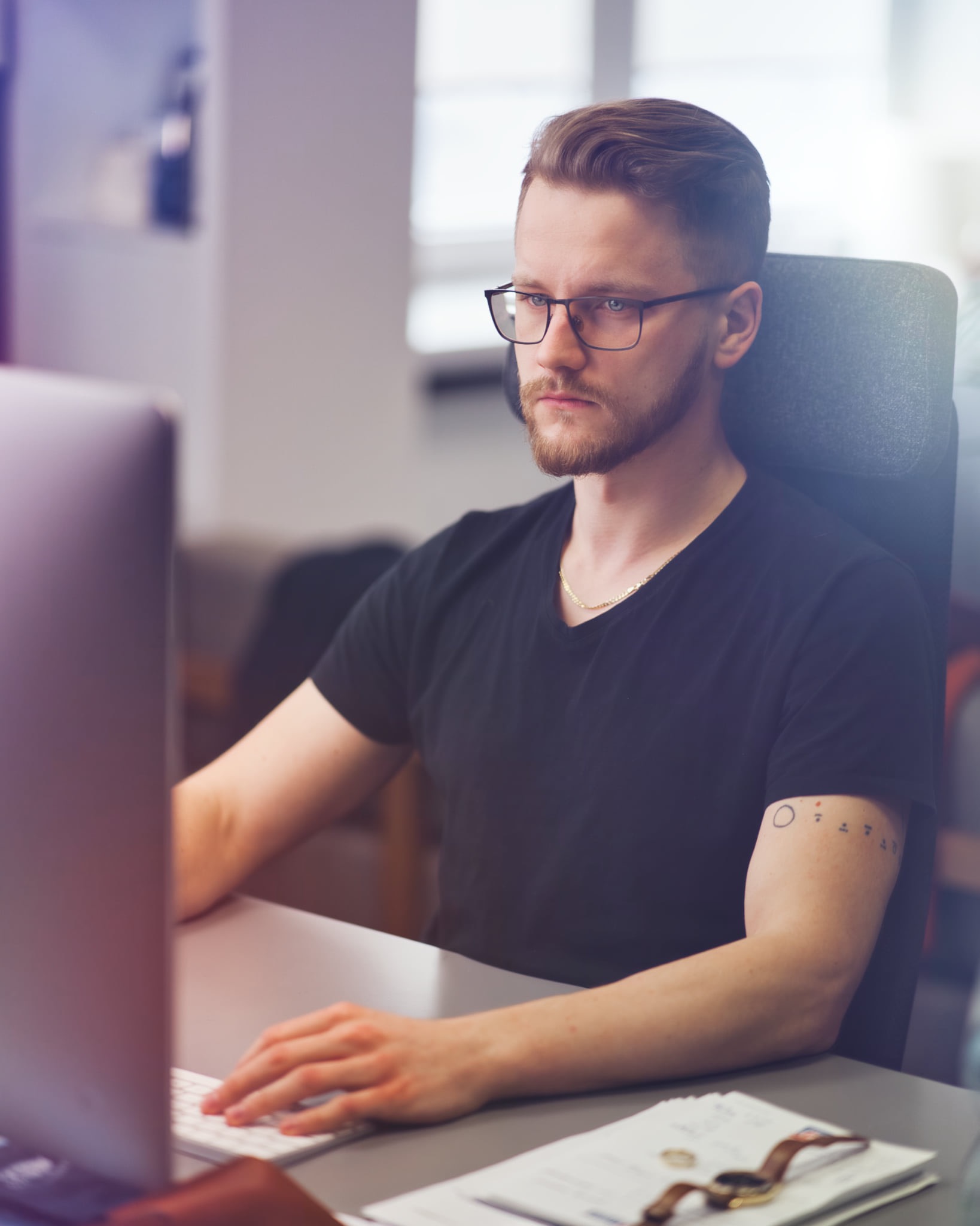 Mężczyzna w okularach, z brodą, w czarnej koszulce, siedzi przy biurku i pisze na klawiaturze komputera; na biurku leżą dokumenty i zegarek na skórzanym pasku.