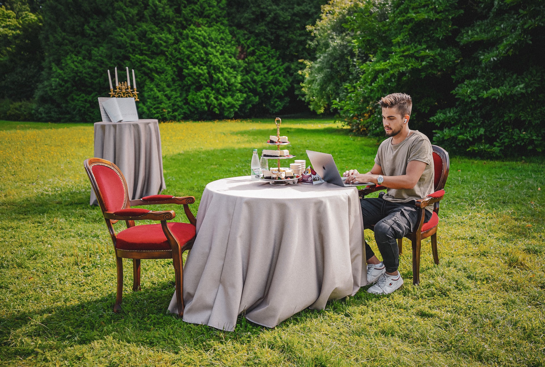 Młody mężczyzna w Rzeszowie pracuje na laptopie przy okrągłym stole na trawie, obok podwieczorek i dekoracje, w tle zieleń drzew.
