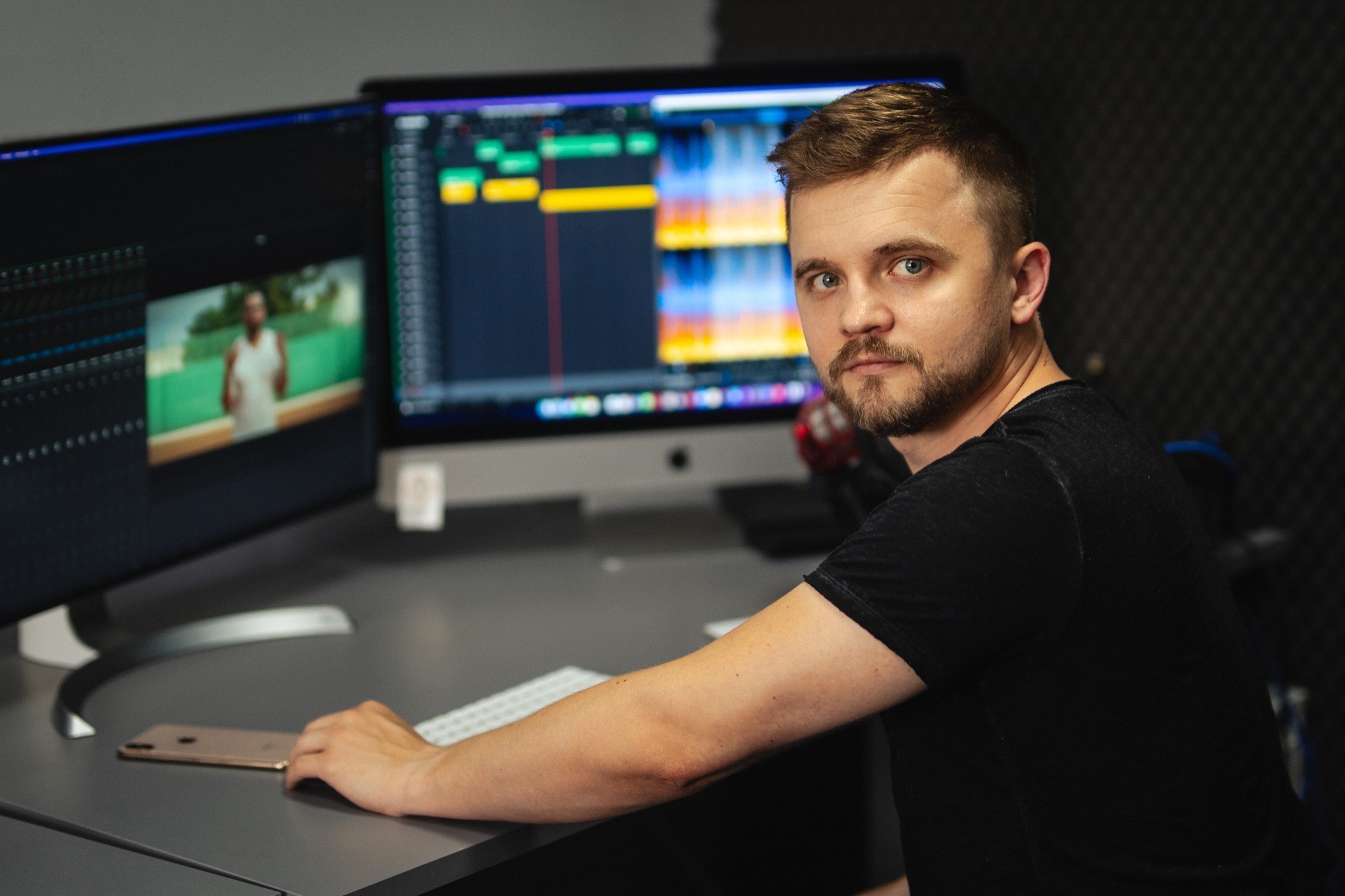 Mężczyzna z brodą siedzi przed dwoma monitorami, edytując wideo z mężczyzną w białej koszulce na tle zieleni i kolorową ścieżką dźwiękową.