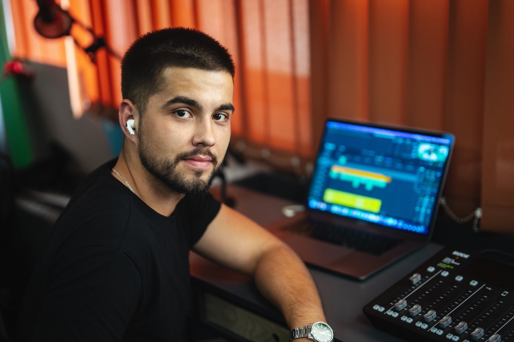 Młody mężczyzna z brodą, w słuchawkach bezprzewodowych, siedzi przed laptopem z otwartym programem do edycji audio i mikserem dźwięku Rode, spogląda w obiektyw.