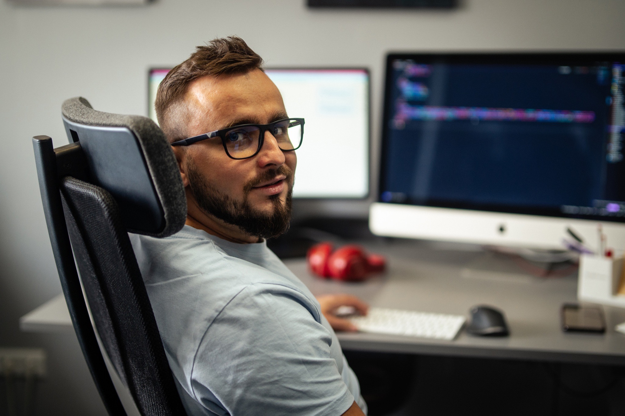 Mężczyzna w okularach siedzi na krześle biurowym, odwrócony w stronę kamery, z dwoma monitorami pokazującymi kod programistyczny w tle.
