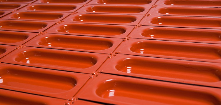 Szereg pomarańczowych, metalowych elementów z podłużnymi wgłębieniami, prawdopodobnie fragmenty obróbek blacharskich, ułożonych równo na płaskiej powierzchni.