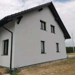 Nowoczesny dom z białą elewacją, czarnym dachem i rynnami, widok z boku, na tle pochmurnego nieba. Widoczne wystające drewniane elementy pod dachem i fragment ogrodu.