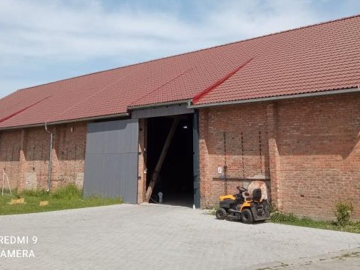 Czerwony dach z dachówki karpiówki na murowanym budynku gospodarczym z otwartymi szarymi wrotami i kosiarką do trawy obok