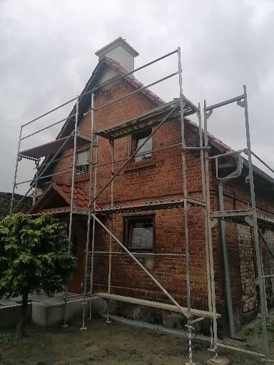 Dom z cegły z rusztowaniem, widoczny fragment dachu z dachówką i komin, elewacja w trakcie prac.