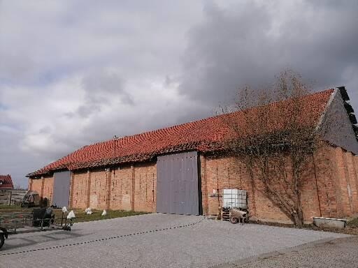Ceglany budynek gospodarczy z czerwonym dachem z dachówki, szarymi wrotami i zbiornikiem na wodę na zewnątrz, widok z niskiej perspektywy w pochmurny dzień.