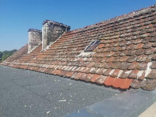 Stary dach z dachówki ceramicznej pokryty mchem i zabrudzeniami, widoczne dwa kominy z cegły oraz okno dachowe, w tle błękitne niebo.