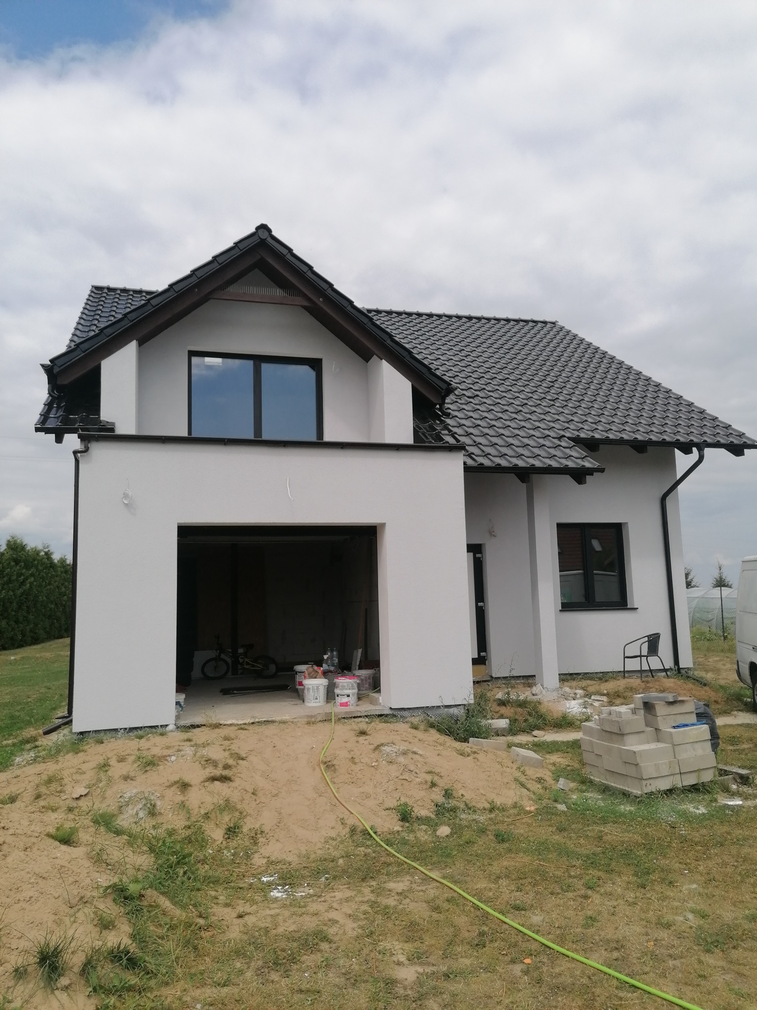 Nowo otynkowany dom jednorodzinny z ciemną dachówką i czarnymi ramami okiennymi, widoczne wiaderka z farbą i rower w otwartym garażu.