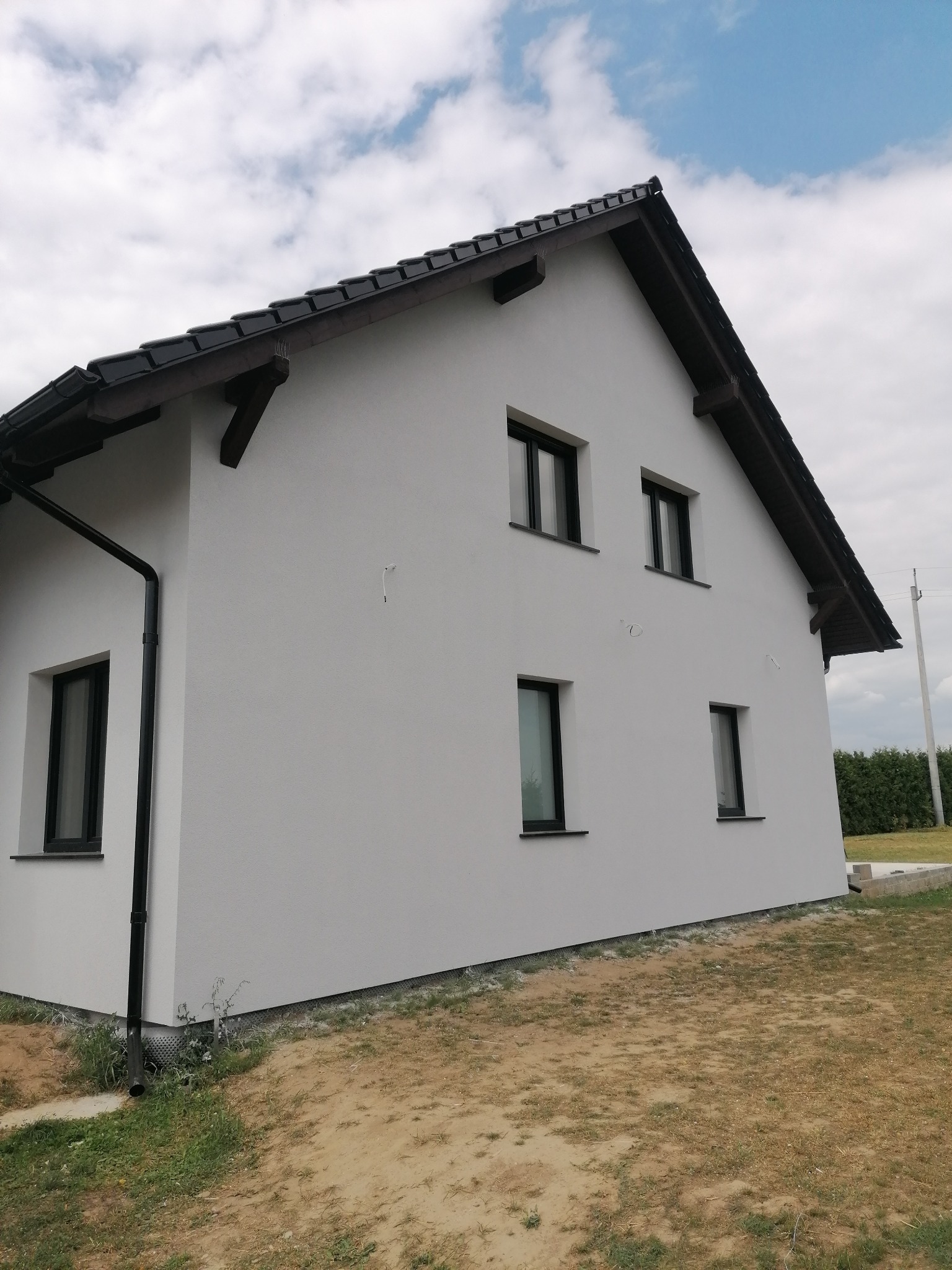 Nowoczesny dom z białą elewacją, czarnym dachem i rynnami, widok z boku, na tle pochmurnego nieba. Widoczne wystające drewniane elementy pod dachem i fragment ogrodu.