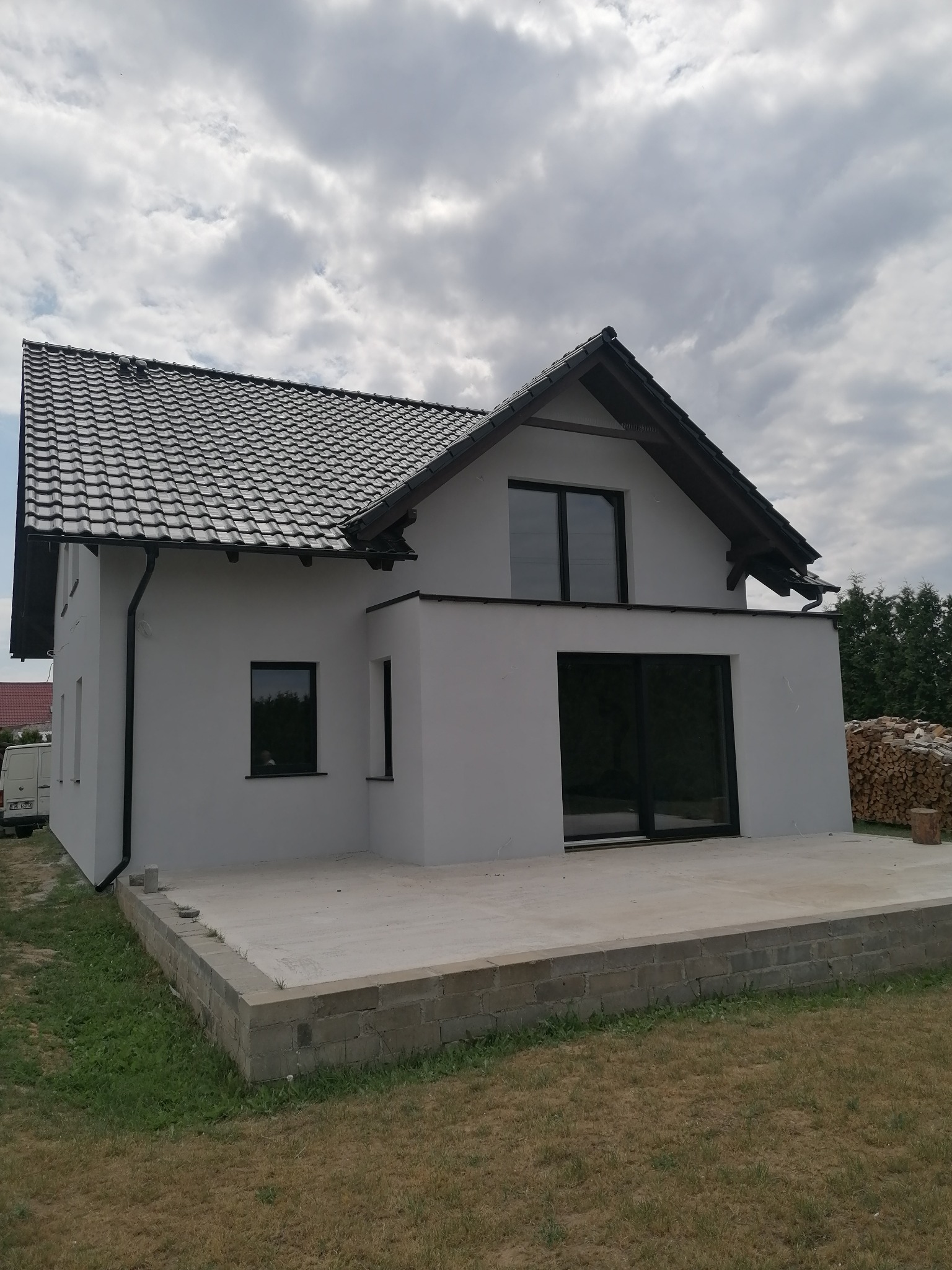Wykończony dom jednorodzinny z szarą dachówką, białymi ścianami i czarnymi ramami okiennymi, widok z zewnątrz na betonowy taras i stos drewna.