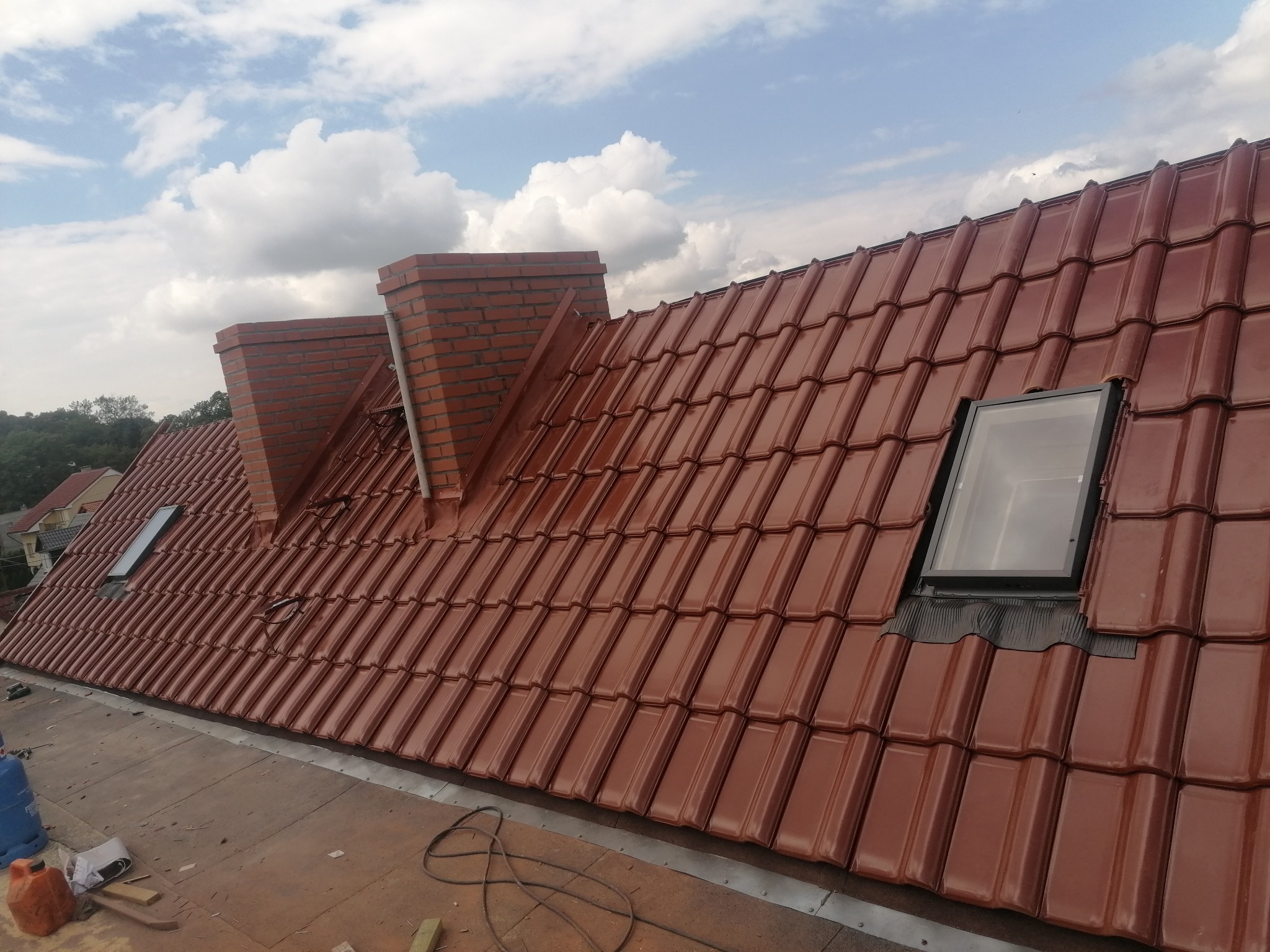 Nowy dach z dachówki ceramicznej w kolorze ceglastym, z dwoma kominami z cegły i oknami dachowymi, widok z góry w słoneczny dzień z chmurami.