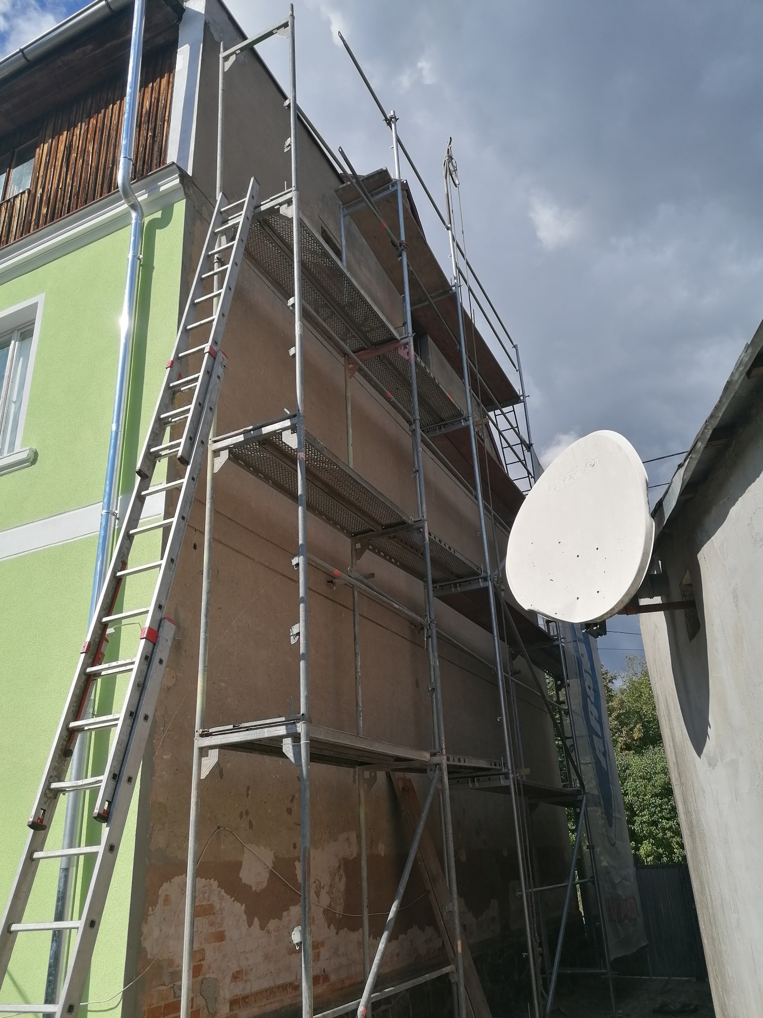 Budynek w trakcie prac elewacyjnych, z widocznym rusztowaniem, drabiną opartą o ścianę i anteną satelitarną na sąsiednim budynku.