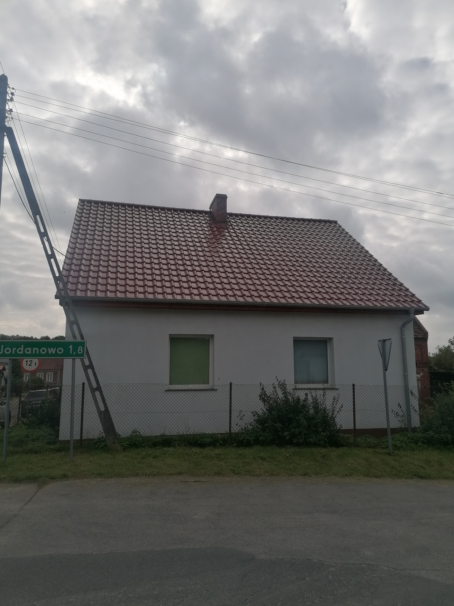 Dom jednorodzinny z nową, czerwoną dachówką ceramiczną, widoczny z poziomu ulicy, z zielonym znakiem drogowym 'Jordanowo 1,8' w kadrze.