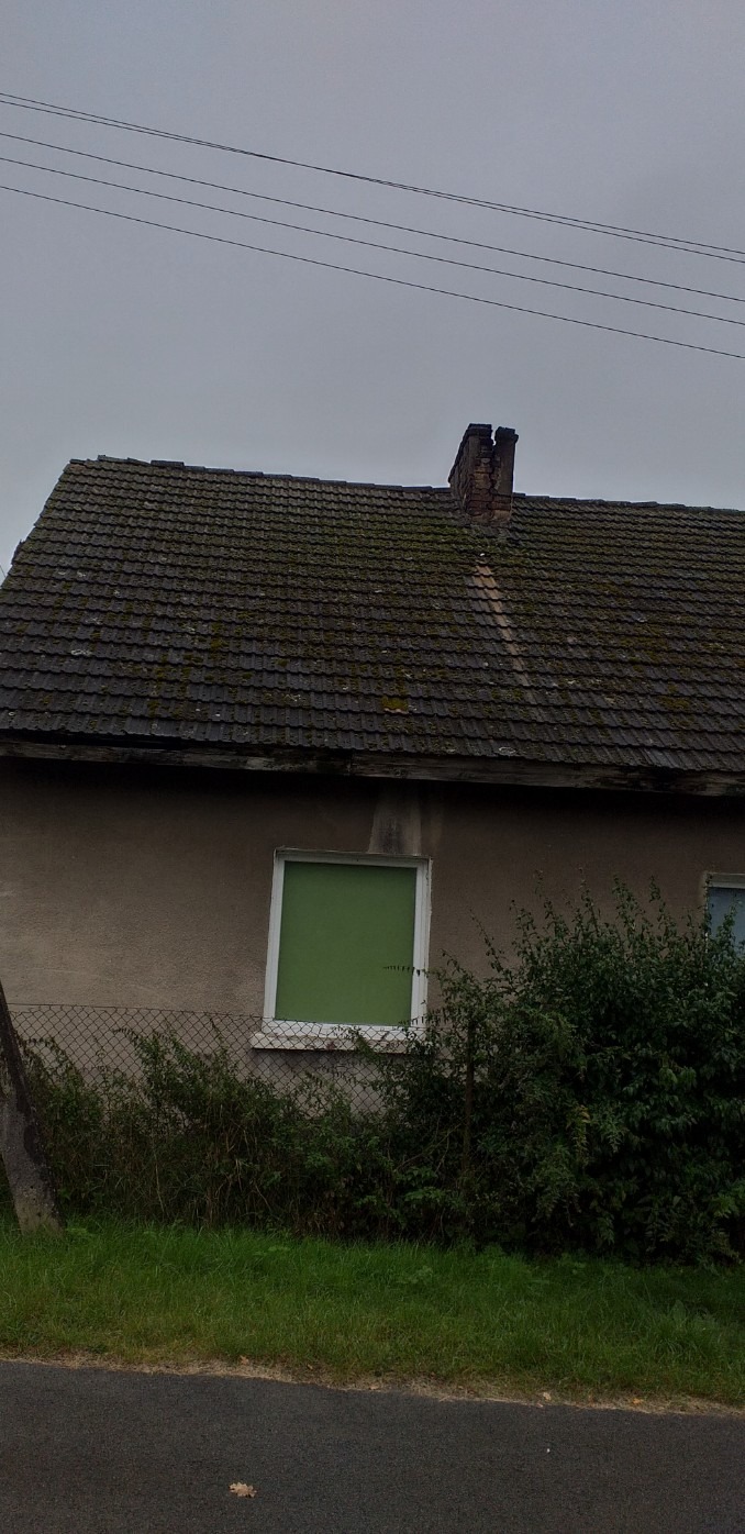 Stary dom z ciemną, zarośniętą mchem dachówką i zniszczonym kominem, widok z poziomu ulicy w pochmurny dzień.