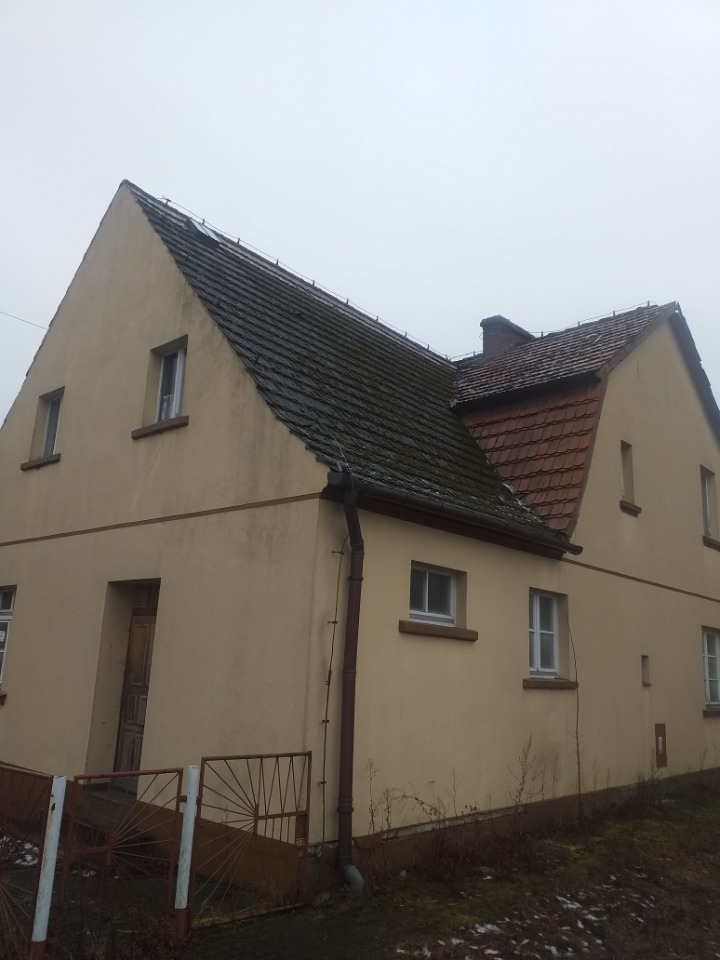 Dwukondygnacyjny dom z beżową elewacją i częściowo zarośniętym, ciemnym dachem, z widoczną rynną spustową po lewej stronie.