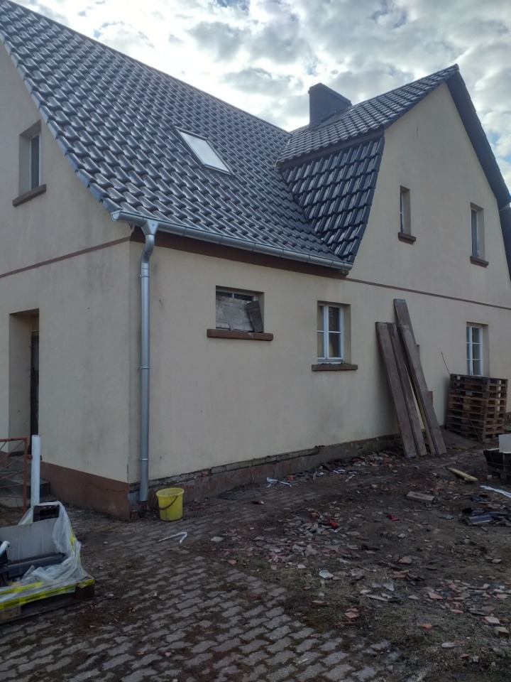 Dwukondygnacyjny budynek z nowym, ciemnoszarym dachem z dachówką, jasnobeżową elewacją i nowymi rynnami. Na ziemi gruz i materiały budowlane, widoczne deski oparte o ścianę i paleta.