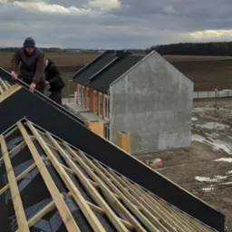 Montaż dachu z widocznym szkieletem więźby, czarną membraną dachową, oraz dwoma dekarzami pracującymi na dachu w pochmurny dzień. W tle surowy, nieotynkowany dom w stanie deweloperskim.