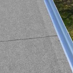 Szczegółowe ujęcie pokrycia dachowego z papy z widoczną linią łączenia i nową, błyszczącą, metalową rynną, z fragmentem zieleni w tle.