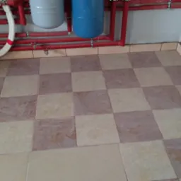 Ułożona podłoga z kwadratowych płytek w odcieniach beżu i brązu, widoczne czerwone rury instalacji grzewczej i białe rury doprowadzające wodę, szara ściana w tle.