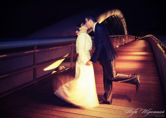 Pocałunek pary młodej na tle oświetlonego mostu nocą, mężczyzna w garniturze unosi nogę w geście radości, suknia ślubna kobiety delikatnie rozmyta ruchem.