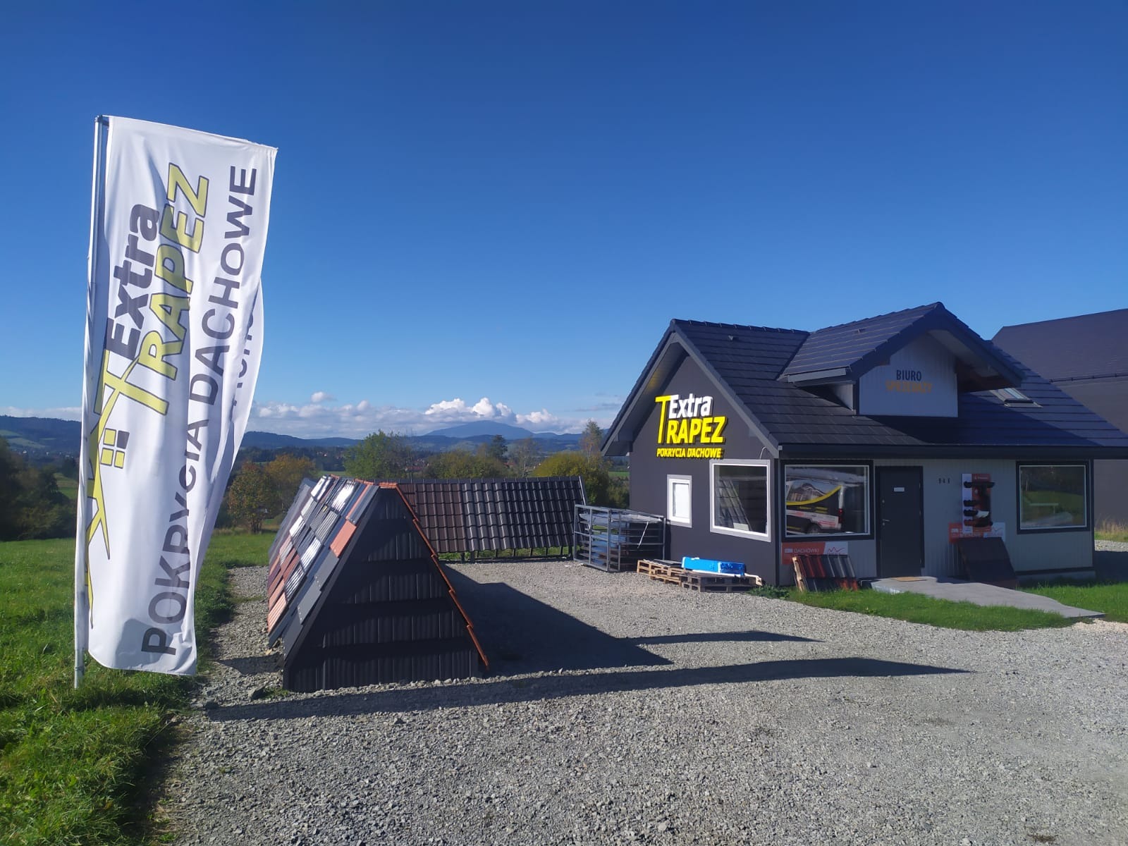 Ekspozycja dachówek i blach trapezowych przed biurem firmy Extra Trapez z widokiem na góry w tle.