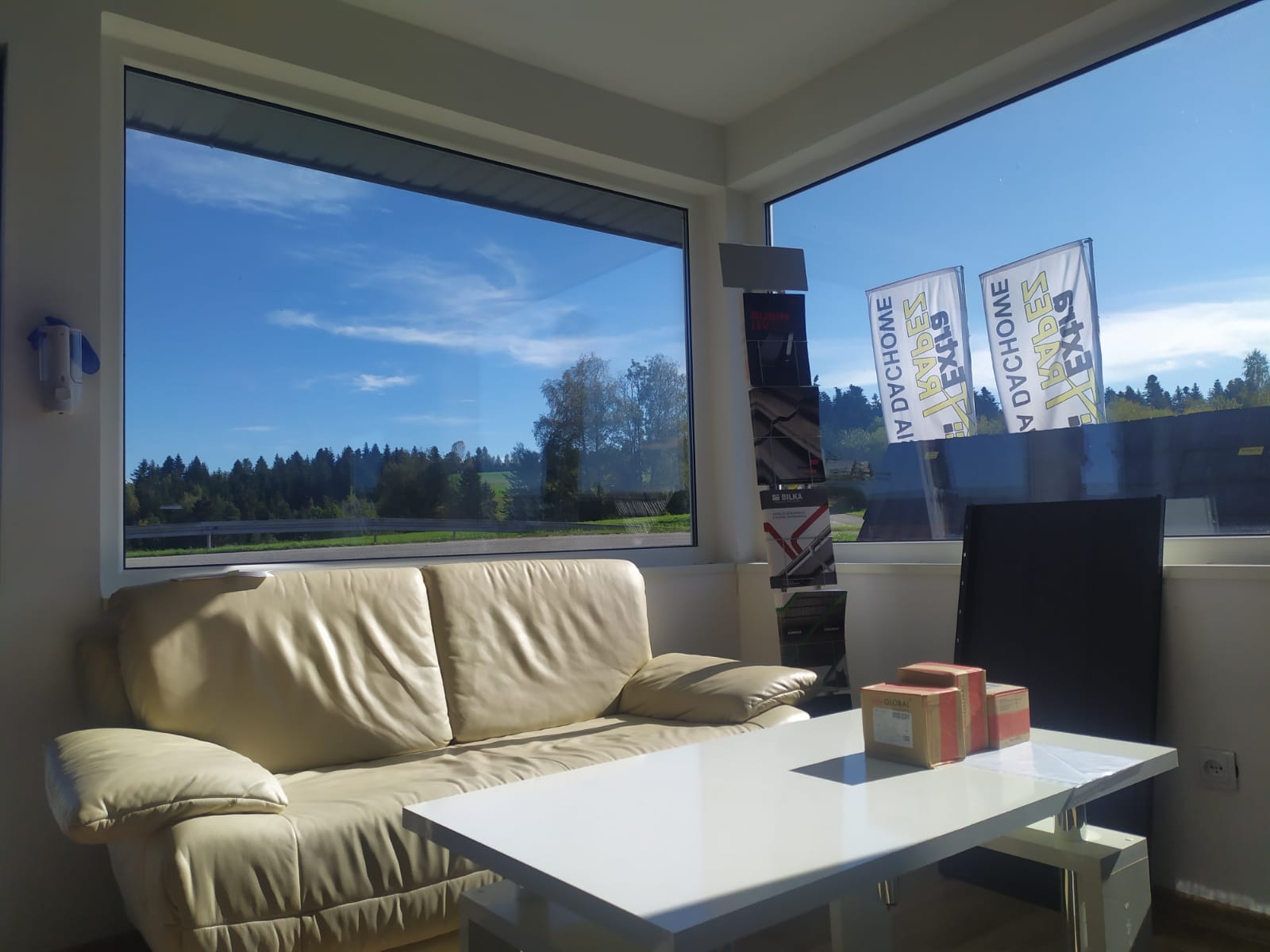 Wnętrze biura z widokiem na krajobraz, sofa, stolik i ekspozycja materiałów budowlanych, widoczne flagi reklamowe producenta pokryć dachowych.