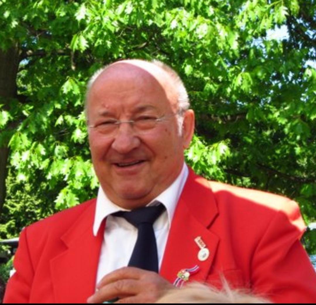 Uśmiechnięty mężczyzna w czerwonej marynarce i okularach, z odznaczeniami, na tle zielonych liści.