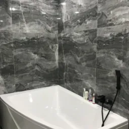 Biała wanna narożna w łazience wyłożonej szarymi płytkami imitującymi marmur, czarna bateria wannowa i prysznicowa.