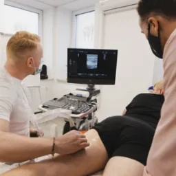 Lekarz w masce wykonuje badanie USG nogi pacjenta, widoczny ekran aparatu USG z obrazem przekroju tkanki, jasne wnętrze gabinetu.