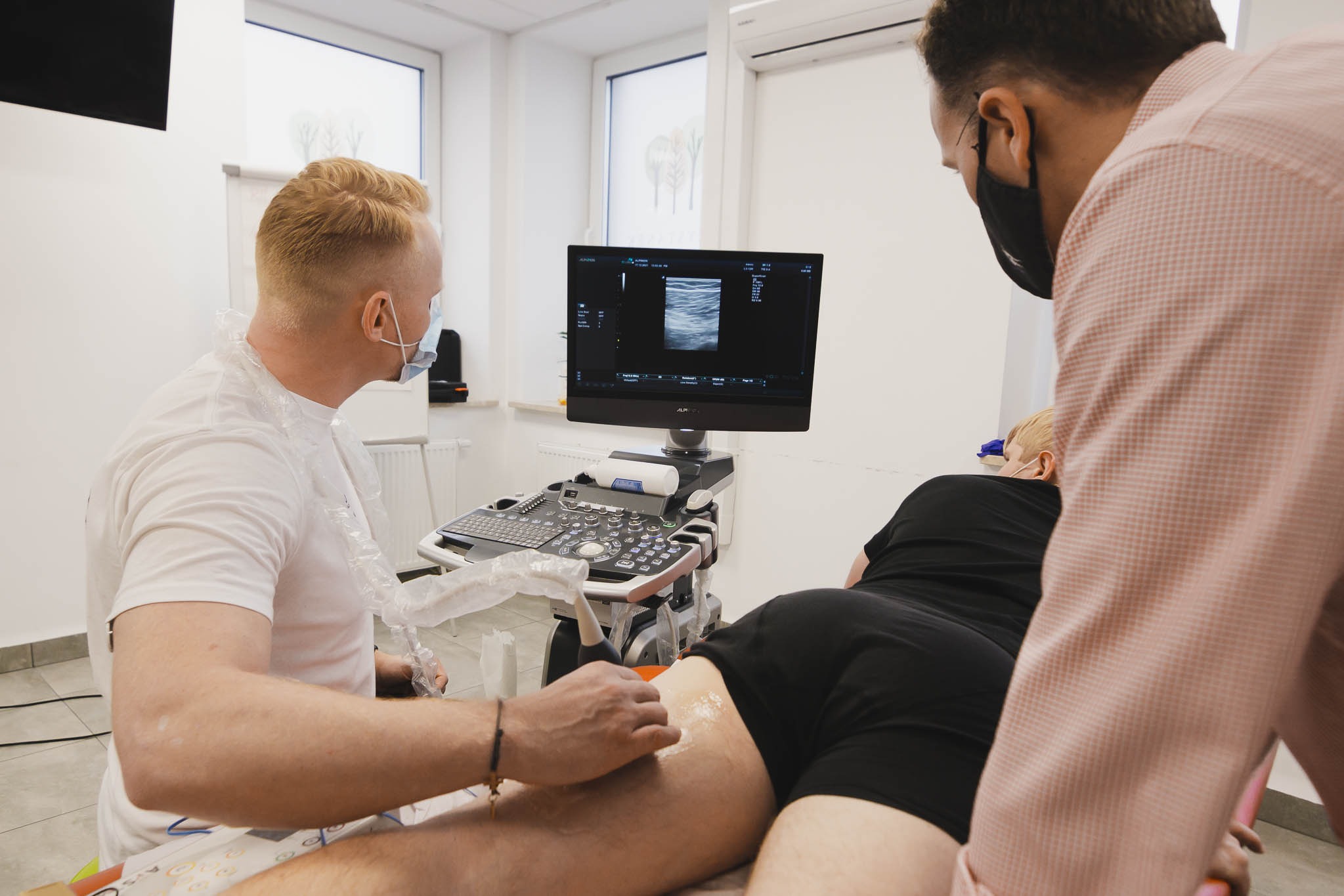Lekarz w masce wykonuje badanie USG nogi pacjenta, widoczny ekran aparatu USG z obrazem przekroju tkanki, jasne wnętrze gabinetu.