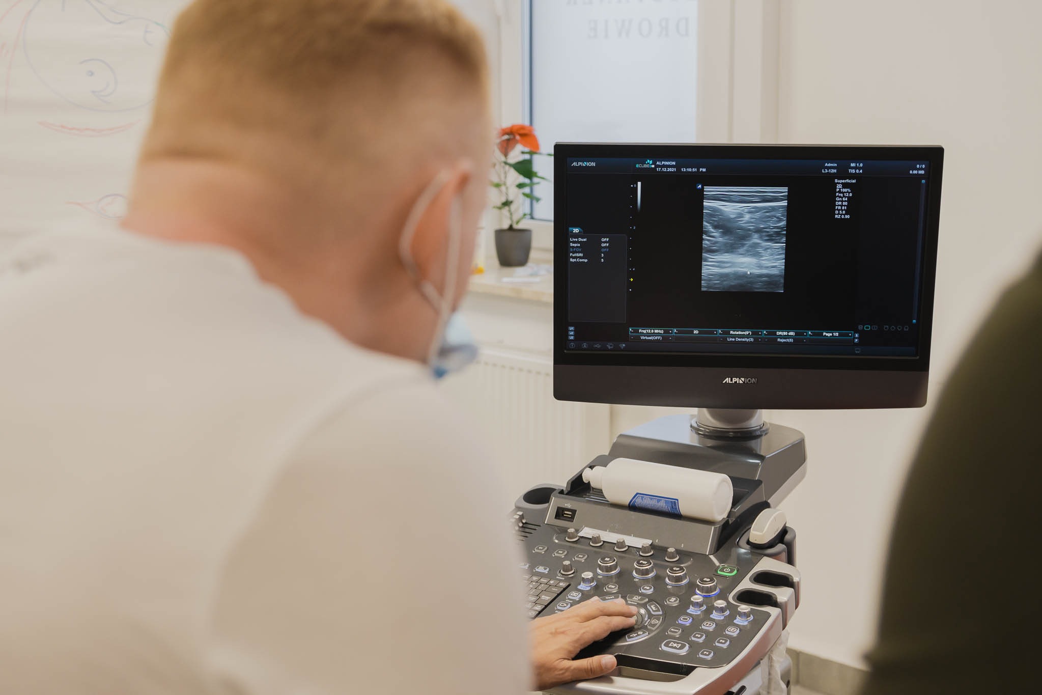 Lekarz w masce obsługuje aparat USG, na ekranie widoczny obraz ultrasonograficzny, obok okno z kwiatem.