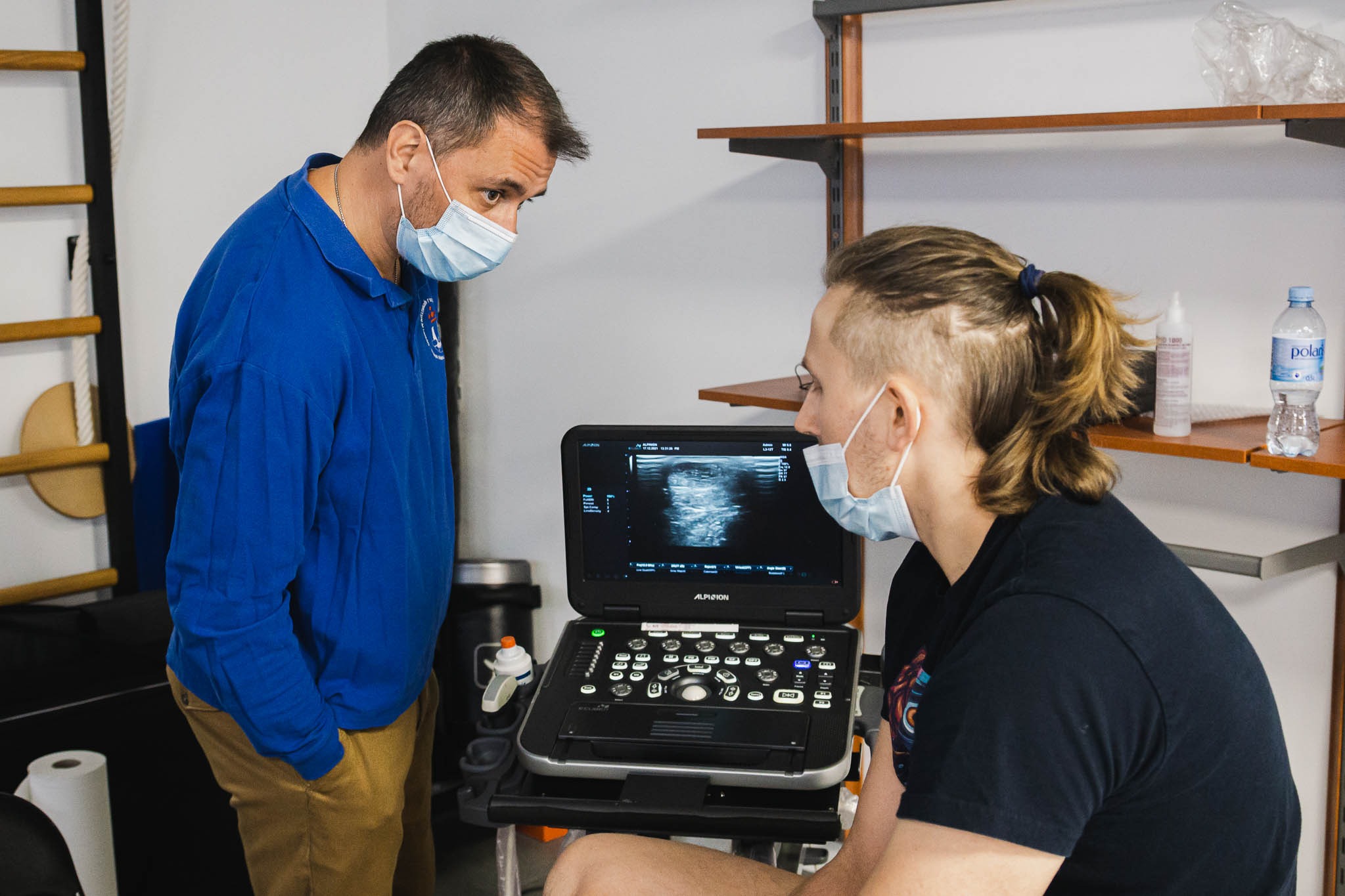 Pacjent w masce podczas badania USG mięśni przez specjalistę w niebieskiej koszuli, widoczny monitor ultrasonografu Alpinion.