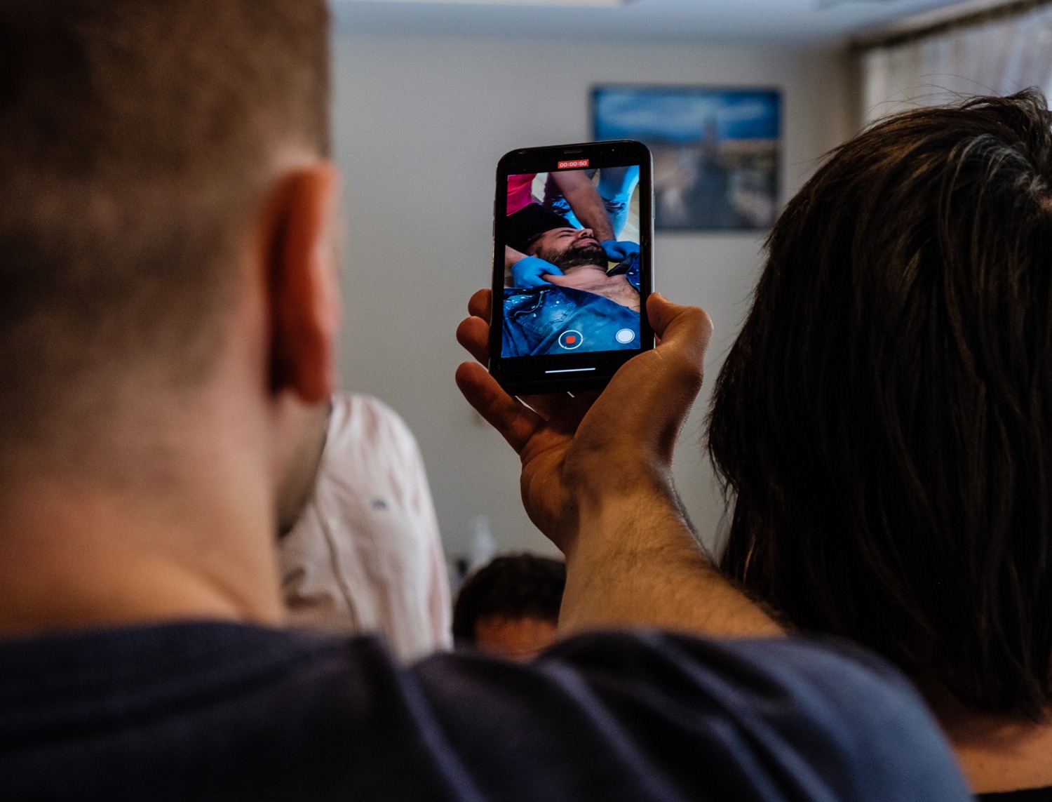Osoba nagrywa telefonem zabieg fizjoterapeutyczny na szyi pacjenta, widoczny ekran telefonu z nagrywanym zabiegiem i fragmenty osób obserwujących.