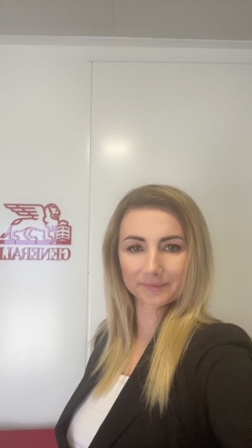 Portret kobiety w czarnej marynarce, z logo firmy ubezpieczeniowej w tle na białej ścianie.