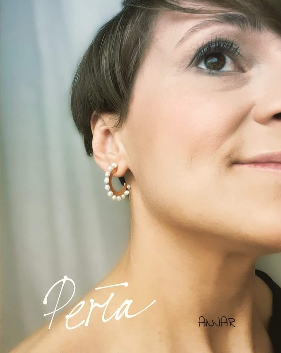 Portret kobiety z kolczykiem w kształcie obręczy z perłami, z widocznym logo marki Perla i Anvar na dekolcie.