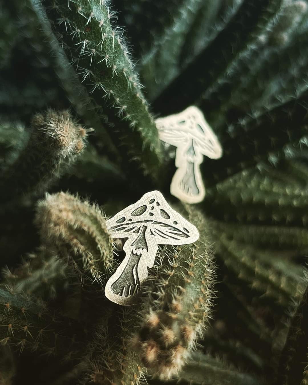 Dwa metalowe znaczki w kształcie grzybów z wygrawerowanymi detalami, umieszczone na kaktusie z długimi, białymi kolcami.