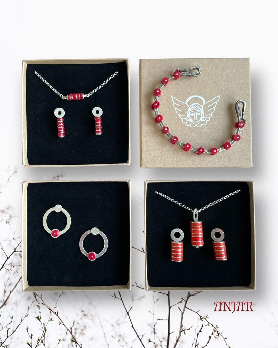 Cztery kwadratowe pudełka z biżuterią: naszyjniki, kolczyki i bransoletka z czerwonymi i srebrnymi elementami, prezentowane na tle z gałązkami.