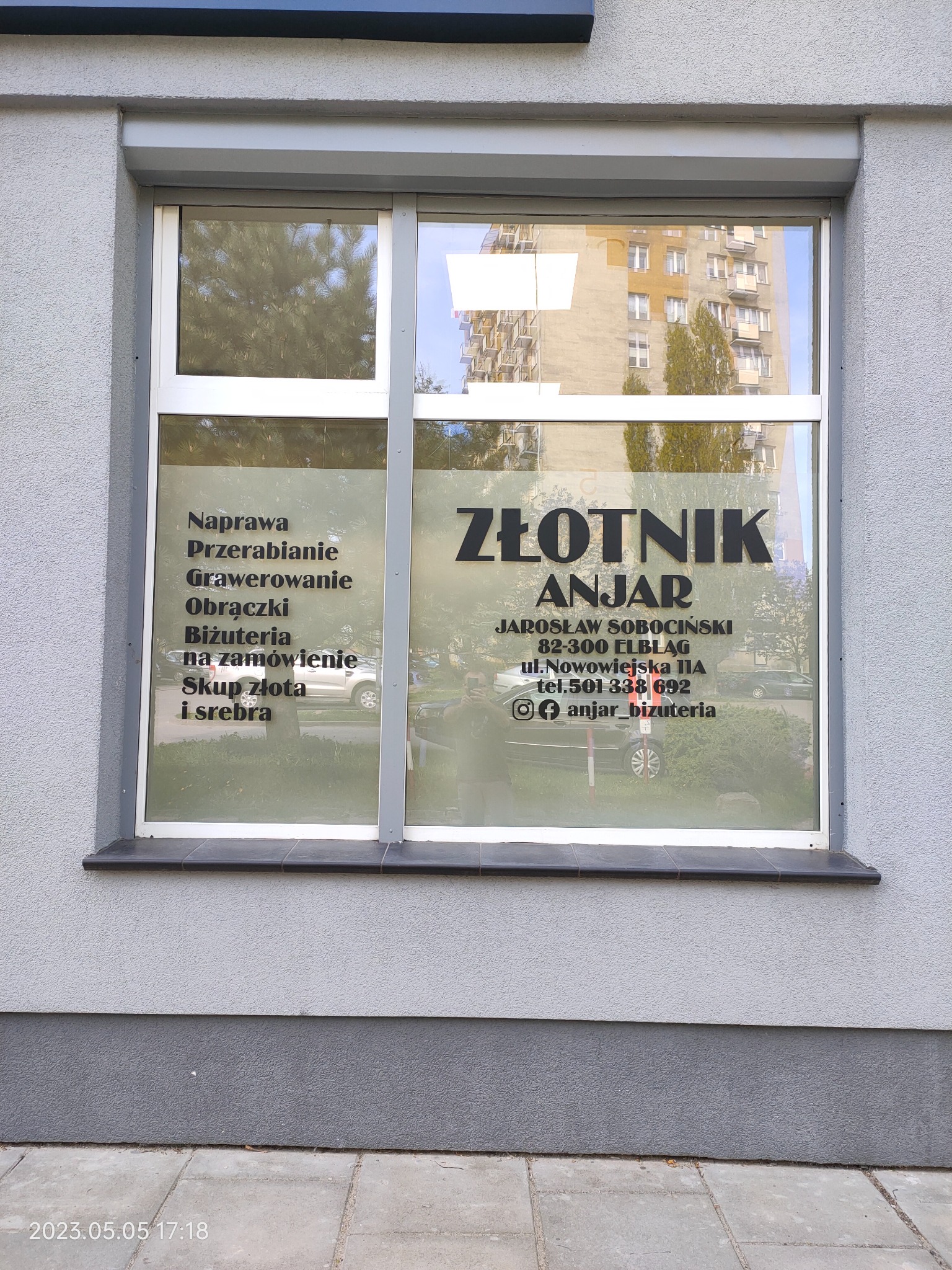 Witryna zakładu złotniczego Anjar z informacjami o naprawie, przerabianiu, grawerowaniu, obrączkach, biżuterii na zamówienie oraz skupie złota i srebra, widoczna ulica i budynek w odbiciu.