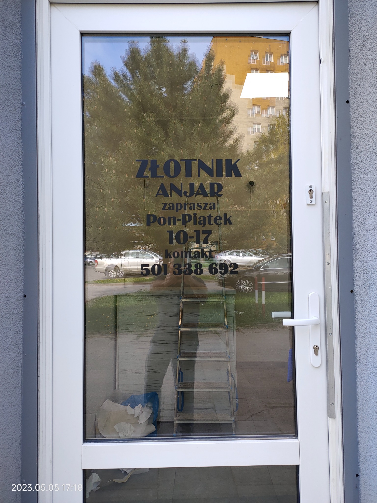 Wizytówka Złotnika Anjar naklejona na szklanych drzwiach wejściowych, widoczne godziny otwarcia i numer telefonu, w tle odbicie drzewa i budynków, w rogu data wykonania zdjęcia.