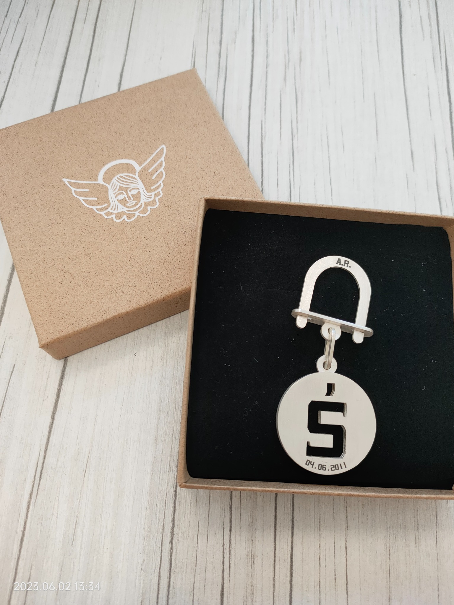 Srebrny breloczek z wygrawerowaną cyfrą '5' i datą '04.06.2011' w pudełku prezentowym z motywem anioła, leżący na drewnianym tle.