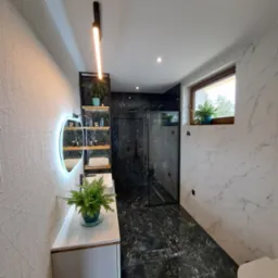 Nowoczesna łazienka z czarno-białymi marmurkowymi płytkami, szklaną kabiną prysznicową, owalnym lustrem z podświetleniem LED, wiszącą szafką umywalkową z paprotką w doniczce i czarnym regałem...