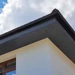 Fragment elewacji budynku z widoczną podbitką dachową w kolorze ciemnego brązu, białą ścianą i drewnianą ramą okienną na tle błękitnego nieba z białymi obłokami.