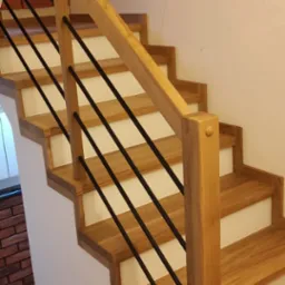 Jasne, dębowe stopnie z białymi podstopnicami i czarnymi, metalowymi balustradami, widok z dołu na tle ściany z cegły.