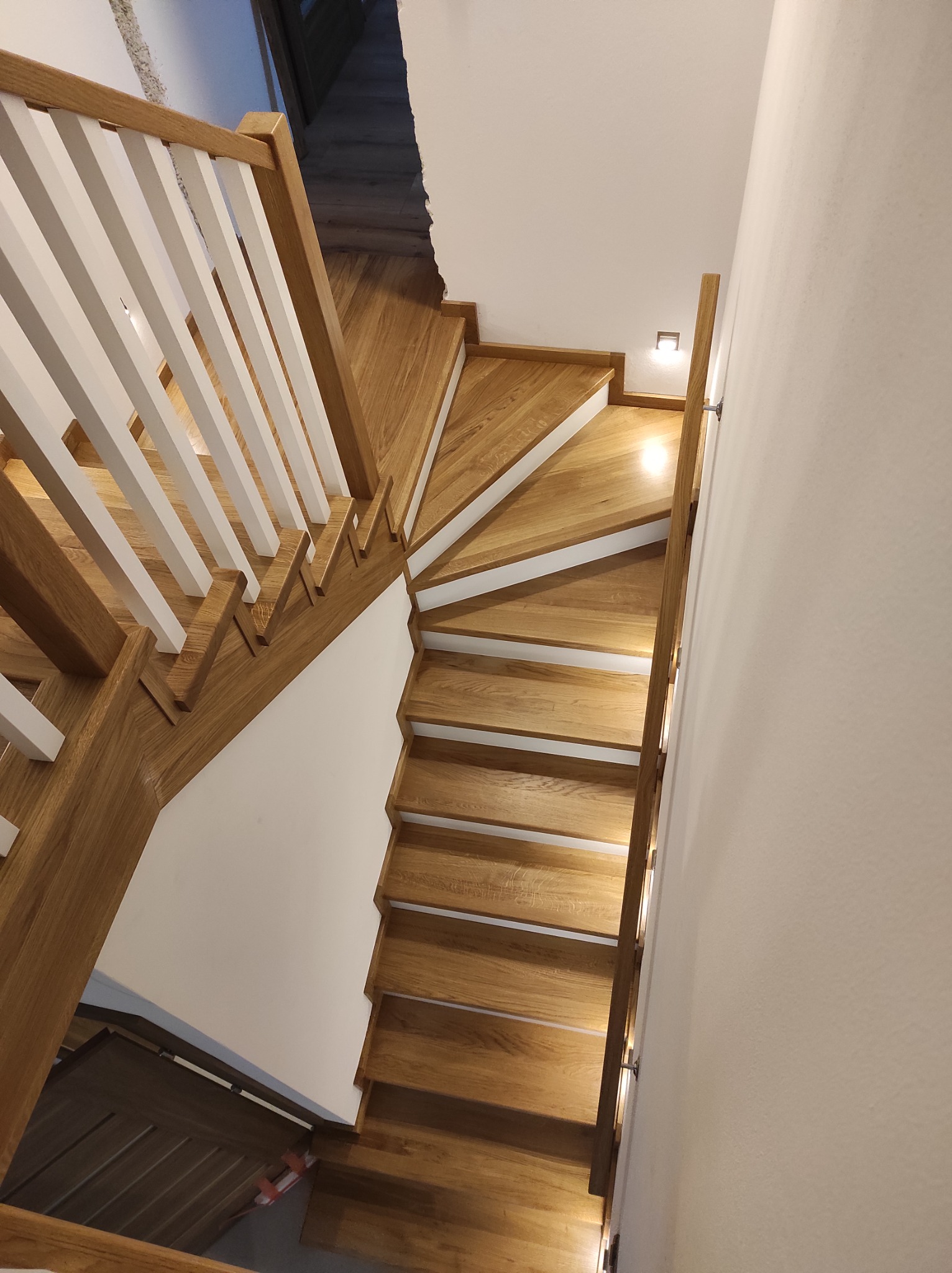 Widok z góry na drewniane schody kręcone z białymi balustradami, oświetlone małymi lampkami ściennymi.